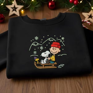 May include: Black crew neck sweatshirt with an embroidered design of Charlie Brown, Snoopy, and Woodstock on a sled in a snowy landscape. The design features a red hat, yellow shirt, and a wooden sled. The design is in a winter theme.