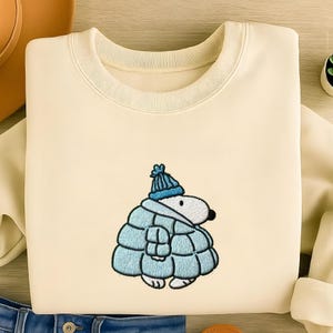 以下が含まれることがあります： クリーム色のスウェットシャツ。青い帽子、スカーフ、コートを着た刺繍された漫画のキャラクターが特徴です。デザインは中央に配置されています。スウェットシャツは柔らかい素材で作られています。