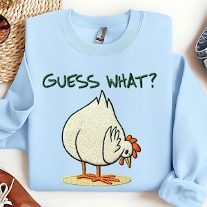 May include: Light blue sweatshirt with the embroidered phrase "GUESS WHAT?" above a cartoon chicken. The chicken is white with orange feet, beak, and comb, standing on a yellow circle.