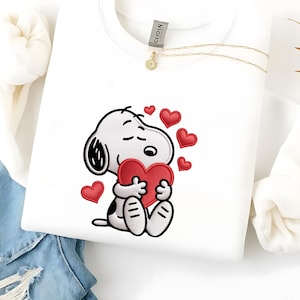 Puede incluir: Sudadera blanca con un diseño bordado de Snoopy sosteniendo un corazón rojo, rodeado de corazones rojos más pequeños. Se ve un collar dorado con un pequeño colgante. La sudadera está doblada sobre una superficie de mezclilla.