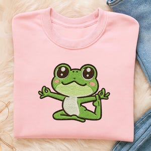 May include: A light pink sweatshirt featuring an embroidered green frog in a yoga pose. The frog has large, expressive eyes and is positioned in a seated asana. The sweatshirt has a crew neck and long sleeves, with a soft, comfortable appearance.