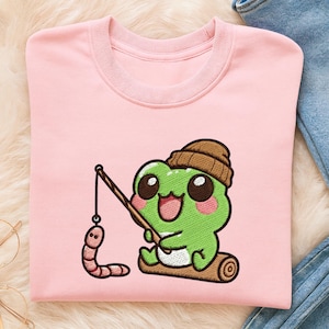 May include: A light pink sweatshirt featuring an embroidered cartoon frog wearing a brown beanie and fishing with a worm. The frog is sitting on a log. The sweatshirt has a round neckline and long sleeves.