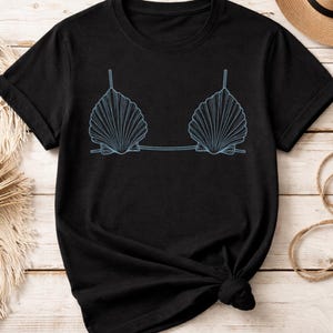 May include: Black t-shirt with a light blue seashell bra design. The graphic shows two stylized seashells joined by a dotted line, with thin straps. The shirt is made of a soft material and has short sleeves.