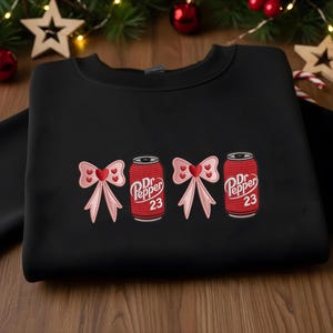May include: Black sweatshirt featuring embroidered designs of pink bows and Dr. Pepper cans. The cans are red with white text and the number 23. The bows have heart-shaped centers.