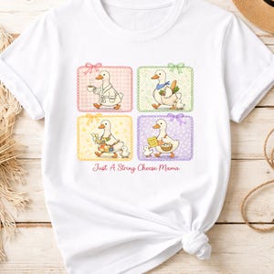 May include: White t-shirt featuring a whimsical design of a duck in four different scenarios, each within a colorful square frame. The duck is depicted with a cup, vegetables, a gift, and a basket. Text reads "Just A String Cheese Mama."