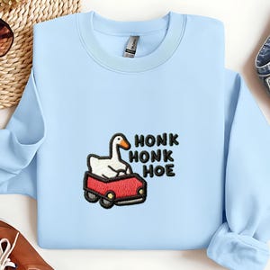 以下が含まれることがあります： 刺繍デザインが施された水色のスウェットシャツ。赤い車に乗った白いガチョウと、黒い文字で「HONK HONK HOE」と書かれています。スウェットシャツはクルーネックで長袖です。