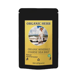 Op de afbeelding: Zwarte zak met een geel etiket met de tekst "ORGANIC HERB" en "ORGANIC MINERALS COARSE SEA SALT". Het etiket toont een persoon die zout oogst. De zak bevat 227 gram grof zeezout.