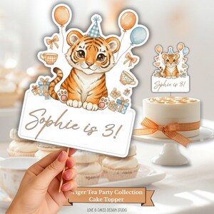 May include: A watercolor-style cake topper featuring a cute tiger cub with balloons and gifts. The sign reads "Sophie is 3!". A small cake with an orange ribbon is also visible, perfect for a birthday celebration.