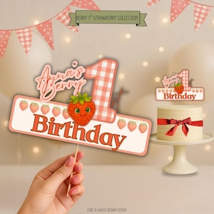 Puede incluir: Una tarta de cumpleaños con una cinta roja y un adorno para tarta que dice "Anna's Berry Birthday". La tarta está sobre un soporte blanco para tartas. La imagen también incluye una pancarta que dice "Berry 1st Strawberry Collection".