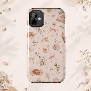 Può includere: Una cover per telefono con sfondo beige chiaro caratterizzata da un motivo ripetuto di fiocchi rosa, croissant su piatti, candele accese, mazzi di fiori, buste, libri e piccoli cuori. Il design include un'apertura per la doppia fotocamera.