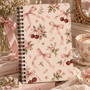 Könnte beinhalten: Ein spiralgebundenes Notizbuch mit cremefarbenem Einband und einem wiederkehrenden Muster aus rosa Schleifen, roten Kirschen und rosa Rosen. Das Design zeigt kleine grüne Blätter und verstreute Blütenblätter sowie eine schwarze Spiralbindung.