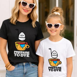 May include: Black and white t-shirts with a graphic of an egg over a cup with the text "PREPARE TO DYE". The egg has a sad face. The cup is yellow, orange, red, and blue. The shirts are worn by two people.