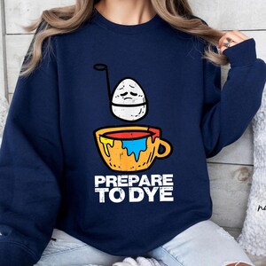 May include: Navy blue sweatshirt with a graphic of an egg with a sad face above a yellow mug filled with paint. The text "PREPARE TO DYE" is printed in white below the mug. The sweatshirt is a casual style.
