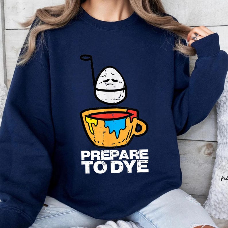 May include: Navy blue sweatshirt with a graphic of an egg with a sad face above a yellow mug filled with paint. The text "PREPARE TO DYE" is printed in white below the mug. The sweatshirt is a casual style.