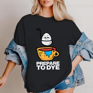 May include: Black t-shirt with a graphic of a sad egg above a cup with colorful paint. The text "PREPARE TO DYE" is printed in white below the cup. The shirt is a casual style.