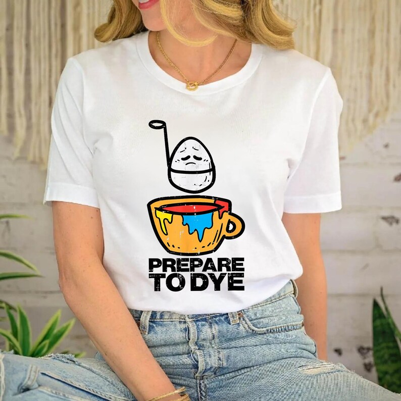 May include: White t-shirt featuring a cartoon egg with a sad face above a yellow mug filled with blue and red paint. The text "PREPARE TO DYE" is printed below the mug in black letters. The shirt is a casual wear item.