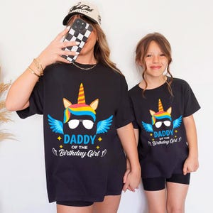 May include: Two black t-shirts with a unicorn graphic and the text "DADDY of the Birthday Girl". The unicorn has a colorful mane, sunglasses, and blue wings. The shirts are short-sleeved.