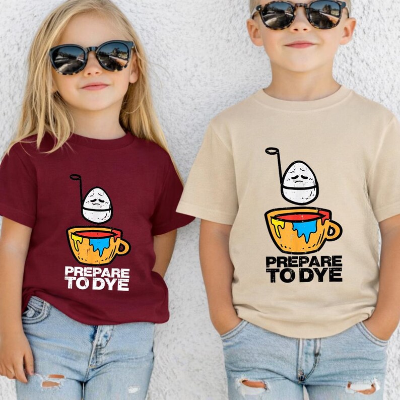 May include: Two children wearing sunglasses and t-shirts. One shirt is maroon, the other is tan. Both shirts feature a cartoon egg over a cup with the text "PREPARE TO DYE" in white.