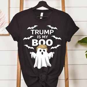 Könnte beinhalten: Schwarzes T-Shirt mit dem weißen Text "TRUMP IS MY BOO", Fledermaus-Silhouetten und einer Geisterillustration mit blonden Haaren. Das T-Shirt wird auf einer Holzleiter präsentiert.
