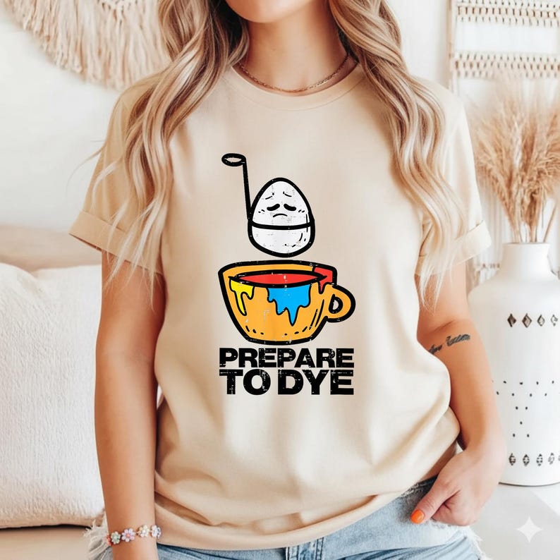 May include: Beige t-shirt featuring a graphic of an egg with a sad face above a colorful cup with the text "PREPARE TO DYE" in black. The design is centered on the front of the shirt.
