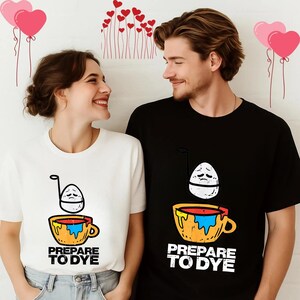 May include: White and black t-shirts with a graphic of an egg in a cup, with the text "PREPARE TO DYE". The egg has a sad face. The cup is yellow and orange with blue and red paint. The shirts are worn by a couple.