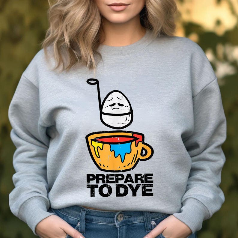 May include: A light gray sweatshirt with a graphic of an egg with a sad face above a yellow cup filled with colorful paint. The text "PREPARE TO DYE" is printed below the cup in black.