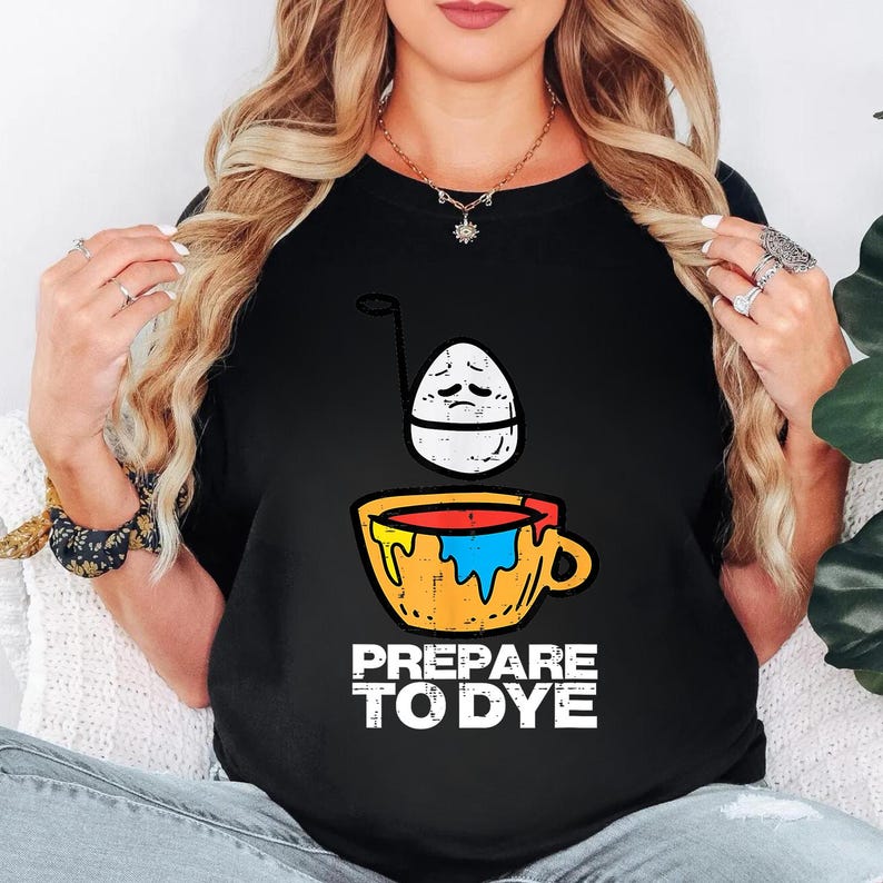 May include: Black t-shirt with a graphic of a sad egg over a cup of dye. The cup is yellow with red, blue, and orange dye. The text "PREPARE TO DYE" is printed in white below the cup.