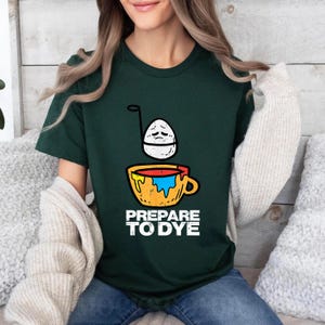 May include: Dark green t-shirt with a graphic of an egg with a sad face above a yellow cup with red and blue paint. The text "PREPARE TO DYE" is printed below the cup in white. The shirt is made of a soft material.