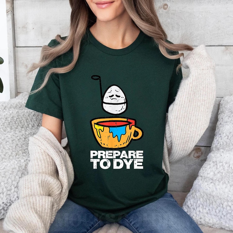 May include: Dark green t-shirt with a graphic of an egg with a sad face above a yellow cup with red and blue paint. The text "PREPARE TO DYE" is printed below the cup in white. The shirt is made of a soft material.