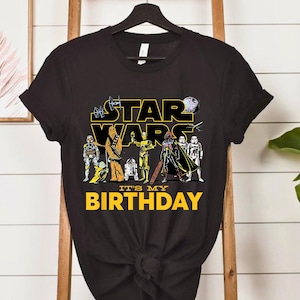 May include: Black t-shirt with a Star Wars graphic, including characters and the yellow text "STAR WARS". Underneath, the yellow text reads "IT'S MY BIRTHDAY". The t-shirt has short sleeves and is made of a soft material.