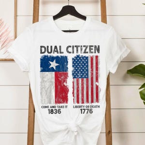 May include: White t-shirt with the text "DUAL CITIZEN" above a graphic of the Texas and U.S. flags. The Texas flag includes a white star on a blue background, and the U.S. flag has red and white stripes. Below the flags are the phrases "COME AND TAKE IT 1836" and "LIBERTY OR DEATH 1776".