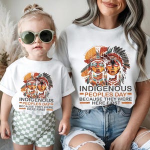 May include: White t-shirts with a graphic featuring two Native American figures in headdresses. The text reads "INDIGENOUS PEOPLES DAY BECAUSE THEY WERE HERE FIRST." One shirt is worn by a child wearing sunglasses, the other by an adult.