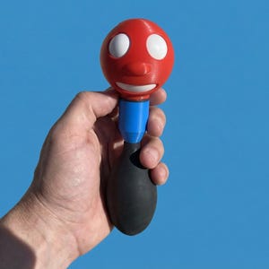 May include: A red and white novelty item with a smiling face design, held against a blue background. The item has a blue cylindrical section and a black bulb at the base. The face has white eyes and a smiling mouth.