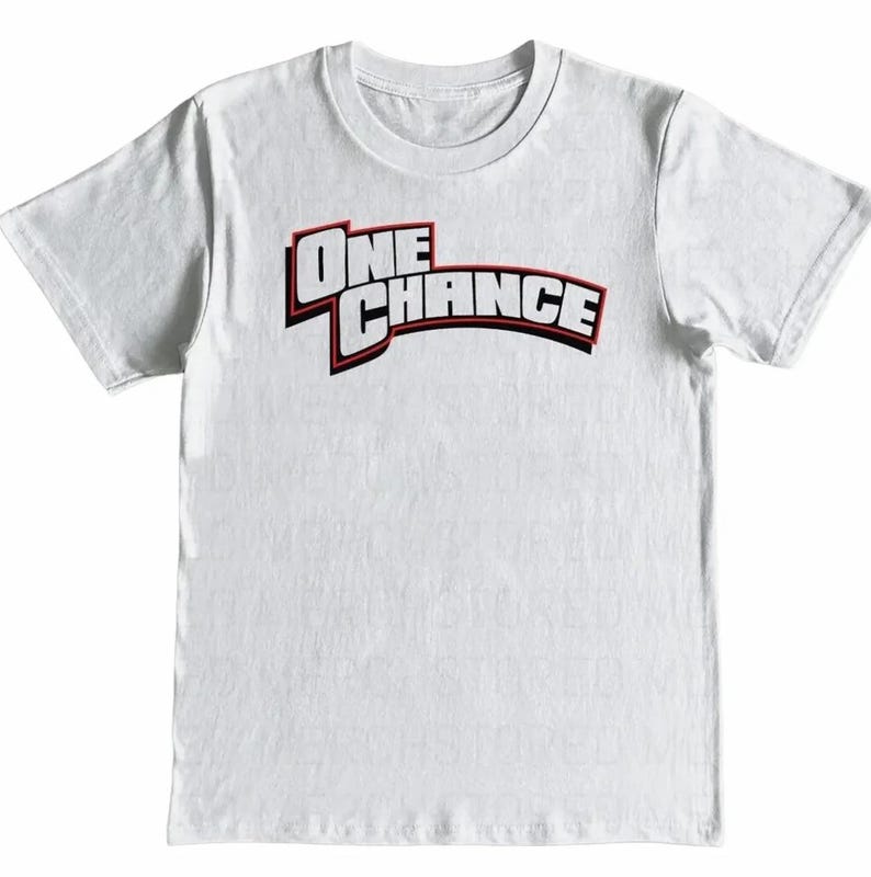 May include: White crew neck t-shirt with the text "ONE CHANCE" in a bold, stylized font. The text is primarily white with a red outline and a black shadow, creating a 3D effect. The shirt is made of a soft, likely cotton material.