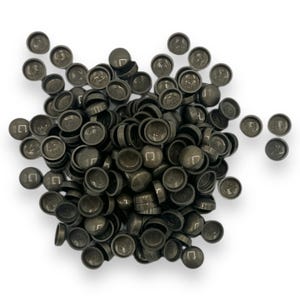 Può includere: Un mucchio di piccoli tappi grigio scuro, dall'aspetto metallico, con un disegno circolare sulla parte superiore. I tappi sono sparsi su uno sfondo bianco, alcuni sovrapposti. I tappi hanno una superficie leggermente riflettente.