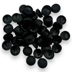 以下が含まれることがあります： 白い背景に散らばる黒いプラスチック製のプラグのコレクション。プラグは円形で、わずかにドーム状の上面とくぼんだ内部があります。一部には目に見えるマーキングがあります。