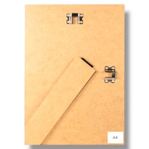 May include: Back of a rectangular picture frame with a light brown wooden backing. The frame has two metal clips at the top and one at the bottom. A cardboard stand is attached to the back. The label "A4" is visible.