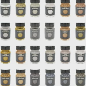May include: A collection of twenty-four small glass jars filled with various metallic-toned paints. Each jar features a black screw-top lid and a black label with the text Liquid Metal printed in white, showcasing the specific paint color inside.