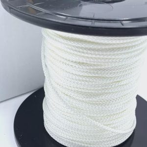 May include: A black plastic reel holding a tightly wound spool of white cord. The cord is made of a woven material. The spool is partially visible against a white background.