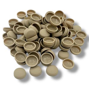 以下が含まれることがあります： ベージュ色のプラスチック製のキャップの山。各キャップは丸みを帯びた上部と円形のベースを備えています。キャップは滑らかでマットな仕上がりで、白い背景に散らばっています。各キャップの内側には「Jap Drum」の文字があります。