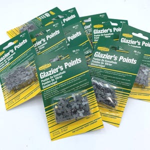 May include: Several packets of Fletcher Glazier's Points are shown. Each packet has a green and yellow label with the product name in multiple languages. The packets contain small, grey, T-shaped glazing points, used for securing glass in frames.