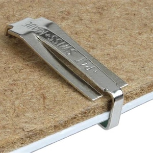 以下が含まれることがあります： 「SWISS MADE」と「PAT」の刻印が入った銀色の金属クリップが、茶色の繊維状の素材に取り付けられています。 クリップは湾曲したデザインで、素材を固定したり、一緒に保持したりするために使用される可能性があります。 背景は明るい茶色です。