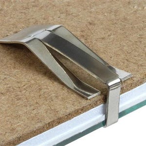 May include: A shiny, silver-colored metal picture frame clip. The clip is designed to hold a picture frame in place. The clip is attached to a brown surface, with a white and green surface visible.