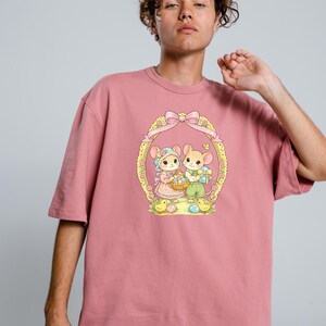 Puede incluir: Camiseta rosa polvoriento con un gr&aacute;fico de Pascua. El dise&ntilde;o muestra dos ratones de dibujos animados dentro de un marco ovalado decorativo, sosteniendo cestas y huevos. El dise&ntilde;o utiliza colores pastel y pollitos.