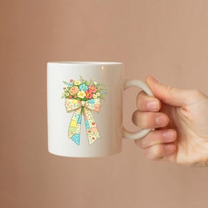 Puede incluir: Taza de cer&aacute;mica blanca con un ramo de flores y un dise&ntilde;o de lazo de patchwork. Las flores son rosas, amarillas, azules y naranjas. El lazo tiene un patr&oacute;n de patchwork con corazones y cuadrados.