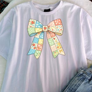 Puede incluir: Camiseta blanca con un gran dise&ntilde;o de lazo colorido. El lazo est&aacute; hecho de cuadrados de patchwork con estampados florales, de fresas y de lunares. El centro del lazo tiene un detalle de fresa. La camiseta se combina con unos vaqueros azules.