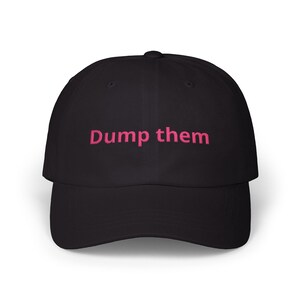 Könnte beinhalten: Eine schwarze Baseballkappe mit dem Schriftzug "Dump them", der in pinkfarbenem Garn auf der Vorderseite eingestickt ist. Die Kappe hat eine strukturierte Krone, einen gebogenen Schirm und einen Knopf oben, was ein schlichtes Design ergibt.