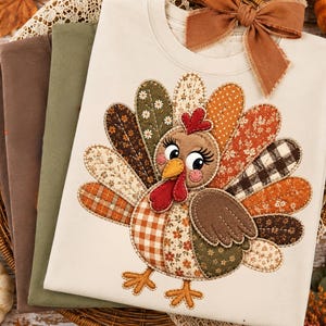 May include: A cream-colored t-shirt featuring a large, colorful turkey appliqué made from various fabric patterns including plaid, floral, and gingham. The turkey has a red wattle, orange beak, and long eyelashes. A brown fabric bow is attached at the neck.