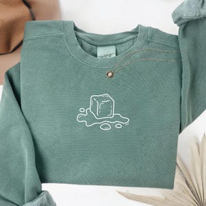 May include: A sage green sweatshirt with a crew neck and long sleeves. A white line drawing of a melting ice cube is centered on the front. A gold necklace with a small pendant is visible. The sleeves are cuffed.