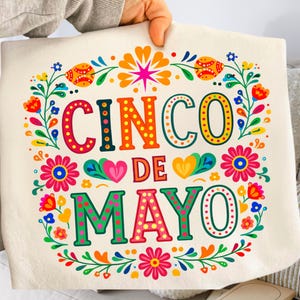 May include: A cream-coloured tote bag featuring the words "CINCO DE MAYO" in bright, decorative lettering. The text is framed by a vibrant floral wreath with red, orange, yellow, and blue accents. The bag is held up by a person.